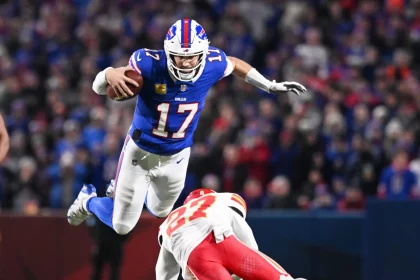 NFL Playoffs: Josh Allen im blauen Trikot mit der Nummer 17 springt über einen Gegenspieler der gegnerischen Mannschaft in Rot und Weiß. Der Spieler hält den Ball fest im Griff, aufgenommen mitten im Flug vor einem verschwommenen Hintergrund begeisterter Fans. Diese Beschreibung wurde mit der FootballR KI automatisch generiert.
