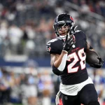 Joe Mixon in schwarz-roter Uniform mit der Nummer 28 läuft mit dem Ball über ein Feld. Im Hintergrund sind unscharfe Zuschauer und ein Schiedsrichter in Streifen zu sehen. Der Fokus liegt auf dem entschlossenen Ausdruck und den Bewegungen des Spielers. Stadiondach verlor Teile. Diese Beschreibung wurde mit der FootballR KI automatisch generiert.