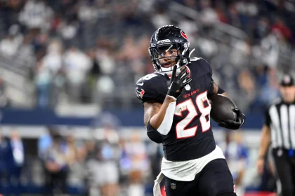 Joe Mixon in schwarz-roter Uniform mit der Nummer 28 läuft mit dem Ball über ein Feld. Im Hintergrund sind unscharfe Zuschauer und ein Schiedsrichter in Streifen zu sehen. Der Fokus liegt auf dem entschlossenen Ausdruck und den Bewegungen des Spielers. Stadiondach verlor Teile. Diese Beschreibung wurde mit der FootballR KI automatisch generiert.