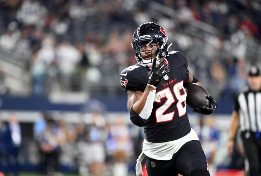 Joe Mixon in schwarz-roter Uniform mit der Nummer 28 läuft mit dem Ball über ein Feld. Im Hintergrund sind unscharfe Zuschauer und ein Schiedsrichter in Streifen zu sehen. Der Fokus liegt auf dem entschlossenen Ausdruck und den Bewegungen des Spielers. Stadiondach verlor Teile. Diese Beschreibung wurde mit der FootballR KI automatisch generiert.