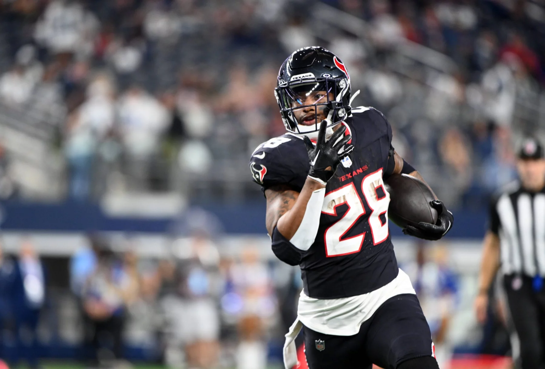 Joe Mixon in schwarz-roter Uniform mit der Nummer 28 läuft mit dem Ball über ein Feld. Im Hintergrund sind unscharfe Zuschauer und ein Schiedsrichter in Streifen zu sehen. Der Fokus liegt auf dem entschlossenen Ausdruck und den Bewegungen des Spielers. Stadiondach verlor Teile. Diese Beschreibung wurde mit der FootballR KI automatisch generiert.