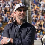 Ein Mann, der Aaron Rodgers ähnelt, mit grauer Kappe, Sonnenbrille und dunkler Jacke, steht lächelnd draußen in einem überfüllten Sportstadion. Er hat Gesichtsbehaarung und trägt eine Uhr am linken Handgelenk. Neben ihm steht ein weiterer Mann mit blauer Kappe und Jacke. Im Hintergrund drängen sich Zuschauer auf den Tribünen. Diese Beschreibung wurde mit der FootballR KI automatisch generiert.