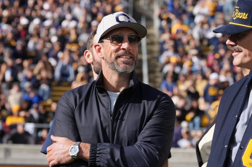 Ein Mann, der Aaron Rodgers ähnelt, mit grauer Kappe, Sonnenbrille und dunkler Jacke, steht lächelnd draußen in einem überfüllten Sportstadion. Er hat Gesichtsbehaarung und trägt eine Uhr am linken Handgelenk. Neben ihm steht ein weiterer Mann mit blauer Kappe und Jacke. Im Hintergrund drängen sich Zuschauer auf den Tribünen. Diese Beschreibung wurde mit der FootballR KI automatisch generiert.