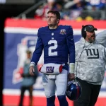 Ein Footballspieler in einem blauen Trikot mit der Nummer 2, Giants-Quarterback Drew Lock, läuft an der Seitenlinie entlang. Er hält einen blauen Helm mit einem roten Streifen in der Hand und geht an Trainern in grauen Kapuzenpullovern mit Camouflage-Muster vorbei. Im Hintergrund weht ein rot-blaues Banner, während Gerüchte über eine Quarterback-Krise geflüstert werden. Diese Beschreibung wurde mit der FootballR KI automatisch generiert.