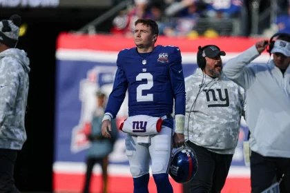 Ein Footballspieler in einem blauen Trikot mit der Nummer 2, Giants-Quarterback Drew Lock, läuft an der Seitenlinie entlang. Er hält einen blauen Helm mit einem roten Streifen in der Hand und geht an Trainern in grauen Kapuzenpullovern mit Camouflage-Muster vorbei. Im Hintergrund weht ein rot-blaues Banner, während Gerüchte über eine Quarterback-Krise geflüstert werden. Diese Beschreibung wurde mit der FootballR KI automatisch generiert.