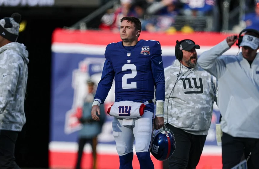 Ein Footballspieler in einem blauen Trikot mit der Nummer 2, Giants-Quarterback Drew Lock, läuft an der Seitenlinie entlang. Er hält einen blauen Helm mit einem roten Streifen in der Hand und geht an Trainern in grauen Kapuzenpullovern mit Camouflage-Muster vorbei. Im Hintergrund weht ein rot-blaues Banner, während Gerüchte über eine Quarterback-Krise geflüstert werden. Diese Beschreibung wurde mit der FootballR KI automatisch generiert.