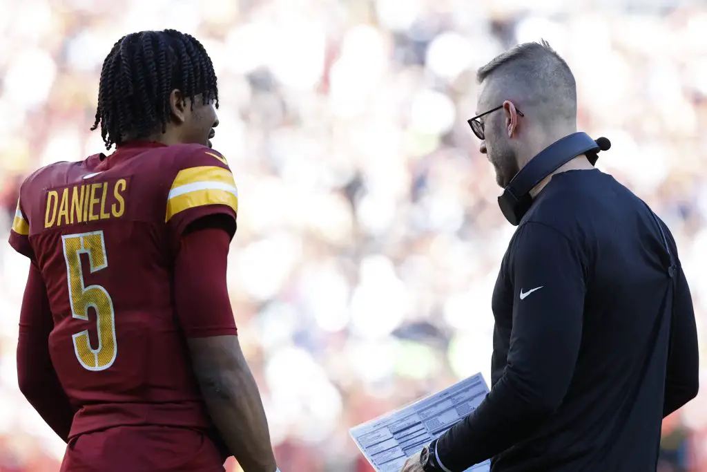 Jayden Daniels in rot-gelber Uniform mit „Daniels“ und der Nummer 5 auf dem Rücken steht auf dem Feld und hört Trainer Kliff Kingsbury aufmerksam zu, der eine Brille und ein schwarzes Langarmshirt trägt und ein Spielblatt in der Hand hält. Im Hintergrund ist eine verschwommene Stadionmenge zu sehen. Diese Beschreibung wurde mit der FootballR KI automatisch generiert.