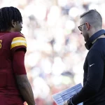 Washington Commanders - Jayden Daniels in rot-gelber Uniform mit „Daniels“ und der Nummer 5 auf dem Rücken steht auf dem Feld und hört Trainer Kliff Kingsbury aufmerksam zu, der eine Brille und ein schwarzes Langarmshirt trägt und ein Spielblatt in der Hand hält. Im Hintergrund ist eine verschwommene Stadionmenge zu sehen. Diese Beschreibung wurde mit der FootballR KI automatisch generiert.