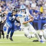 NFL Playoff Picture: Jared Goff in weißer Uniform läuft mit dem Ball und versucht, einem Tackling eines Gegners in dunkler Uniform auszuweichen. Ein weiterer Spieler in Weiß liegt rechts auf dem Boden. Links ist ein Schiedsrichter zu sehen. Im Hintergrund ist eine Zuschauermenge in einem Stadion zu sehen. Diese Beschreibung wurde mit der FootballR KI automatisch generiert.