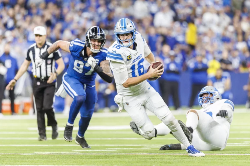 NFL Playoff Picture: Jared Goff in weißer Uniform läuft mit dem Ball und versucht, einem Tackling eines Gegners in dunkler Uniform auszuweichen. Ein weiterer Spieler in Weiß liegt rechts auf dem Boden. Links ist ein Schiedsrichter zu sehen. Im Hintergrund ist eine Zuschauermenge in einem Stadion zu sehen. Diese Beschreibung wurde mit der FootballR KI automatisch generiert.