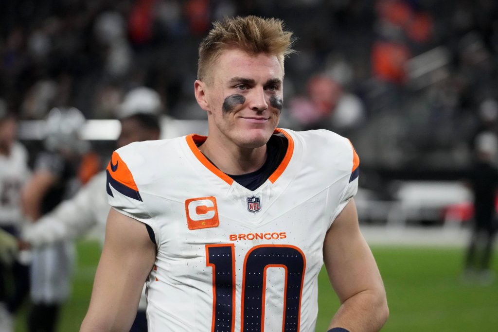 Bo Nix trägt ein weißes Denver Broncos-Trikot mit der Nummer 10 und einem Kapitänsabzeichen, während er auf dem Feld steht. Schwarze Augenschminke betont seinen konzentrierten Blick unter kurzen, zerzausten Haaren. Im verschwommenen Hintergrund sind undeutliche Figuren und Stadionsitze zu sehen. Diese Beschreibung wurde mit der FootballR KI automatisch generiert.