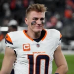 Bo Nix trägt ein weißes Denver Broncos-Trikot mit der Nummer 10 und einem Kapitänsabzeichen, während er auf dem Feld steht. Schwarze Augenschminke betont seinen konzentrierten Blick unter kurzen, zerzausten Haaren. Im verschwommenen Hintergrund sind undeutliche Figuren und Stadionsitze zu sehen. Diese Beschreibung wurde mit der FootballR KI automatisch generiert.