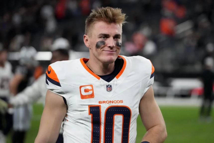 Bo Nix trägt ein weißes Denver Broncos-Trikot mit der Nummer 10 und einem Kapitänsabzeichen, während er auf dem Feld steht. Schwarze Augenschminke betont seinen konzentrierten Blick unter kurzen, zerzausten Haaren. Im verschwommenen Hintergrund sind undeutliche Figuren und Stadionsitze zu sehen. Diese Beschreibung wurde mit der FootballR KI automatisch generiert.
