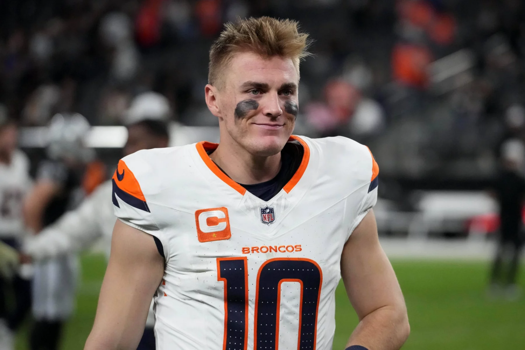 Bo Nix trägt ein weißes Denver Broncos-Trikot mit der Nummer 10 und einem Kapitänsabzeichen, während er auf dem Feld steht. Schwarze Augenschminke betont seinen konzentrierten Blick unter kurzen, zerzausten Haaren. Im verschwommenen Hintergrund sind undeutliche Figuren und Stadionsitze zu sehen. Diese Beschreibung wurde mit der FootballR KI automatisch generiert.