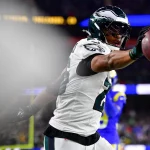 NFL Running Backs: Saquon Barkley in schwarz-weißer Uniform, der einen Helm mit einem Adler-Logo trägt, streckt seinen rechten Arm aus und hält einen Football. Auf dem Feld, mit verschwommenen Zuschauern und blau-gelben Uniformen im Hintergrund, feiert er freudig einen scheinbaren Touchdown. Diese Beschreibung wurde mit der FootballR KI automatisch generiert.