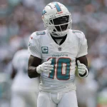 Tyreek Hill, Spieler der Miami Dolphins, steht in weißer Uniform mit der Nummer 10 und dem Kapitänsabzeichen auf dem Footballfeld. Er trägt Helm und Handschuhe und wirkt konzentriert und einsatzbereit, während die verschwommene Menge im Stadion aufmerksam zusieht. Diese Beschreibung wurde mit der FootballR KI automatisch generiert.