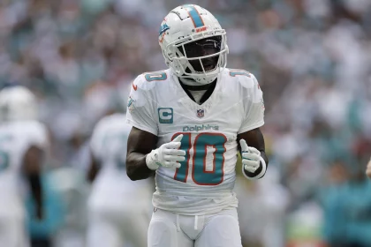 Tyreek Hill, Spieler der Miami Dolphins, steht in weißer Uniform mit der Nummer 10 und dem Kapitänsabzeichen auf dem Footballfeld. Er trägt Helm und Handschuhe und wirkt konzentriert und einsatzbereit, während die verschwommene Menge im Stadion aufmerksam zusieht. Diese Beschreibung wurde mit der FootballR KI automatisch generiert.