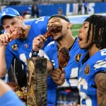 Footballspieler in blauen Trikots, bekannt als die Detroit Lions, feiern, indem sie große Putenkeulen essen. Sie stehen zusammen in einem Stadion, im Vordergrund glänzt eine Trophäe. Ihre Mienen sind freudig, sie genießen den Moment nach einem aufregenden Spiel offensichtlich. Diese Beschreibung wurde mit der FootballR KI automatisch generiert.
