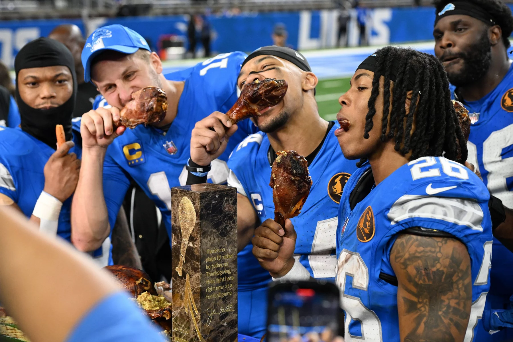 Footballspieler in blauen Trikots, bekannt als die Detroit Lions, feiern, indem sie große Putenkeulen essen. Sie stehen zusammen in einem Stadion, im Vordergrund glänzt eine Trophäe. Ihre Mienen sind freudig, sie genießen den Moment nach einem aufregenden Spiel offensichtlich. Diese Beschreibung wurde mit der FootballR KI automatisch generiert.