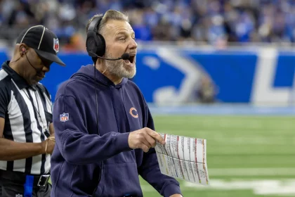 Ein Footballtrainer, der Matt Eberflus von den Bears sehr ähnlich sieht, trägt einen blauen Kapuzenpulli und ein Headset und schreit leidenschaftlich, während er an der Seitenlinie während einer gefühlten Thanksgiving-Niederlage eine Spieltabelle in der Hand hält. Ein Schiedsrichter in einem gestreiften Hemd steht daneben, während das Feld grün und in Vorfreude getaucht bleibt. Diese Beschreibung wurde mit der FootballR KI automatisch generiert.