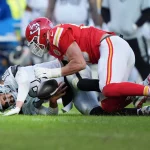 Ein Footballspieler in einem roten Trikot attackiert einen anderen Spieler in Weiß auf einem Rasenfeld. Der attackierte Spieler hält den Football fest, während er auf dem Boden liegt. Die Trikots der Spieler zeigen kontrastierende Teamfarben und im Hintergrund sind mehrere Teamkollegen und Zuschauer zu sehen. Diese Beschreibung wurde mit der FootballR KI automatisch generiert.