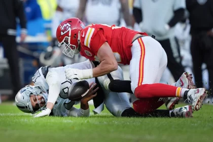Ein Footballspieler in einem roten Trikot attackiert einen anderen Spieler in Weiß auf einem Rasenfeld. Der attackierte Spieler hält den Football fest, während er auf dem Boden liegt. Die Trikots der Spieler zeigen kontrastierende Teamfarben und im Hintergrund sind mehrere Teamkollegen und Zuschauer zu sehen. Diese Beschreibung wurde mit der FootballR KI automatisch generiert.
