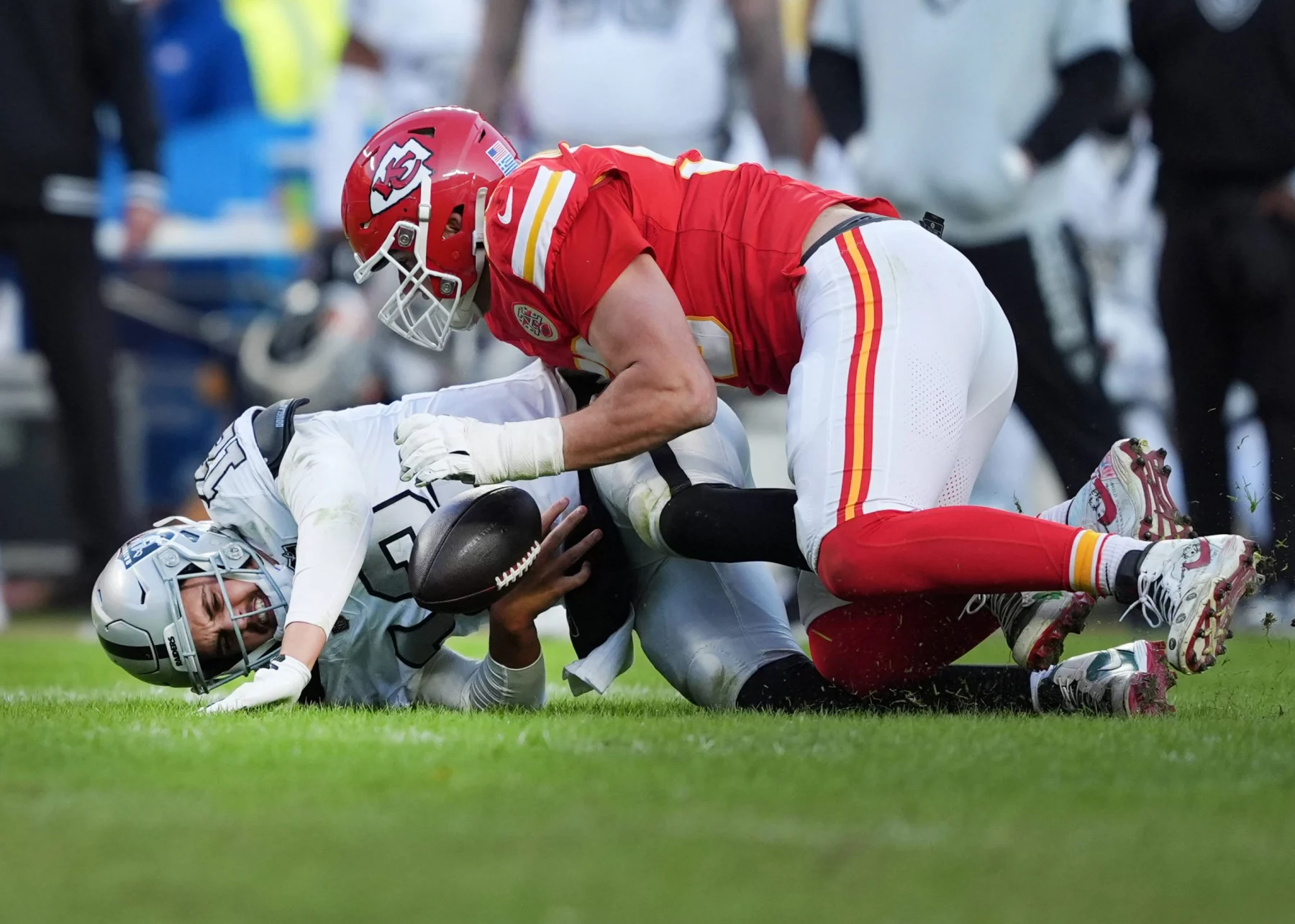 Ein Footballspieler in einem roten Trikot attackiert einen anderen Spieler in Weiß auf einem Rasenfeld. Der attackierte Spieler hält den Football fest, während er auf dem Boden liegt. Die Trikots der Spieler zeigen kontrastierende Teamfarben und im Hintergrund sind mehrere Teamkollegen und Zuschauer zu sehen. Diese Beschreibung wurde mit der FootballR KI automatisch generiert.
