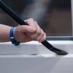 NFL warnt. Eine Hand mit einem blauen Armband benutzt ein Brecheisen, um die Unterseite eines weißen Fensterrahmens vorsichtig aufzubrechen und dabei sicherzustellen, dass kein Schaden entsteht. Diese Beschreibung wurde mit der FootballR KI automatisch generiert.