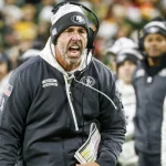 Kyle Shanahan in einer schwarzen San Francisco 49ers Jacke und mit Kopfhörern schreit während eines Footballspiels leidenschaftlich am Spielfeldrand. Er hält ein gefaltetes Blatt mit Spielzügen in der Hand. Im Hintergrund sind Menschen in weißen Jacken verschwommen, was auf einen kalten Tag schließen lässt. Das Stadion ist überfüllt und die Energie ist intensiv. Diese Beschreibung wurde mit der FootballR KI automatisch generiert.