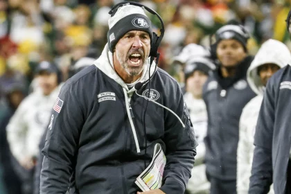 Kyle Shanahan in einer schwarzen San Francisco 49ers Jacke und mit Kopfhörern schreit während eines Footballspiels leidenschaftlich am Spielfeldrand. Er hält ein gefaltetes Blatt mit Spielzügen in der Hand. Im Hintergrund sind Menschen in weißen Jacken verschwommen, was auf einen kalten Tag schließen lässt. Das Stadion ist überfüllt und die Energie ist intensiv. Diese Beschreibung wurde mit der FootballR KI automatisch generiert.