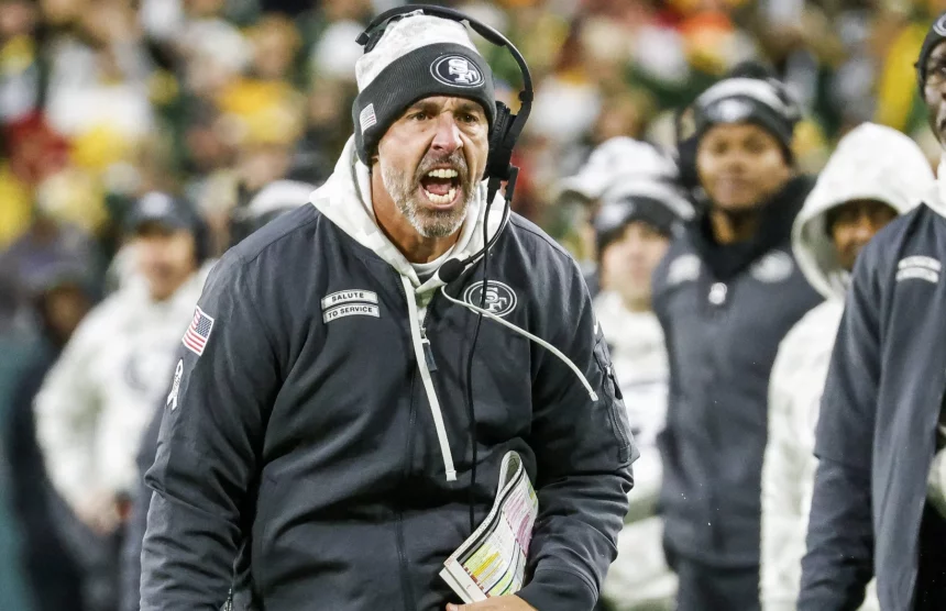 Kyle Shanahan in einer schwarzen San Francisco 49ers Jacke und mit Kopfhörern schreit während eines Footballspiels leidenschaftlich am Spielfeldrand. Er hält ein gefaltetes Blatt mit Spielzügen in der Hand. Im Hintergrund sind Menschen in weißen Jacken verschwommen, was auf einen kalten Tag schließen lässt. Das Stadion ist überfüllt und die Energie ist intensiv. Diese Beschreibung wurde mit der FootballR KI automatisch generiert.