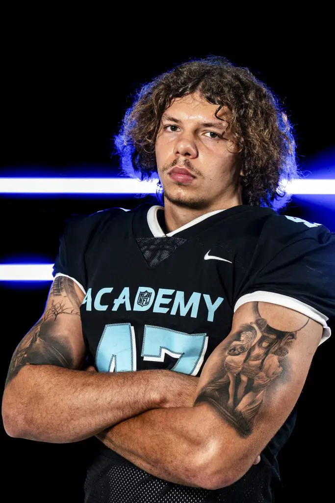 Ein Footballspieler steht selbstbewusst mit verschränkten Armen da. Er trägt ein schwarzes Trikot mit „ACADEMY“ auf der Brust und der Nummer 47. Seine lockigen Haare reichen ihm bis zu den Schultern und er hat Tattoos auf den Armen. Der Hintergrund ist dunkel mit zwei horizontalen blauen Lichtern. Diese Beschreibung wurde mit der FootballR KI automatisch generiert.