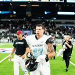 Kevin Bentin in hellblau-weißer Uniform mit der Nummer 47 läuft mit seinem Helm in der Hand über ein Feld. Hinter ihm sind Menschen in schwarzen „ACADEMY“-Trikots, Schiedsrichter und eine Menschenmenge zu sehen. Das Stadion ist hell erleuchtet, was den grünen Rasen und den konzentrierten Gesichtsausdruck des Spielers hervorhebt. Diese Beschreibung wurde mit der FootballR KI automatisch generiert.