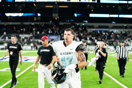 Kevin Bentin in hellblau-weißer Uniform mit der Nummer 47 läuft mit seinem Helm in der Hand über ein Feld. Hinter ihm sind Menschen in schwarzen „ACADEMY“-Trikots, Schiedsrichter und eine Menschenmenge zu sehen. Das Stadion ist hell erleuchtet, was den grünen Rasen und den konzentrierten Gesichtsausdruck des Spielers hervorhebt. Diese Beschreibung wurde mit der FootballR KI automatisch generiert.