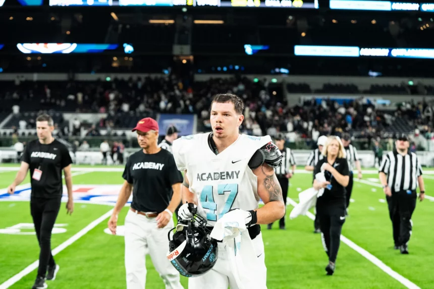 Kevin Bentin in hellblau-weißer Uniform mit der Nummer 47 läuft mit seinem Helm in der Hand über ein Feld. Hinter ihm sind Menschen in schwarzen „ACADEMY“-Trikots, Schiedsrichter und eine Menschenmenge zu sehen. Das Stadion ist hell erleuchtet, was den grünen Rasen und den konzentrierten Gesichtsausdruck des Spielers hervorhebt. Diese Beschreibung wurde mit der FootballR KI automatisch generiert.