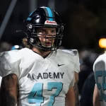 Auf dem Spielfeld steht Kevin Bentin mit schwarzem Helm und weißem Trikot mit der Nummer 47 und „ACADEMY“ auf der Vorderseite. Unter dem Helm sind lange Haare des Spielers zu sehen. Ein anderer Spieler, teilweise zu sehen, trägt ein Trikot mit der Nummer 55. Der Hintergrund ist schwach beleuchtet. Diese Beschreibung wurde mit der FootballR KI automatisch generiert.