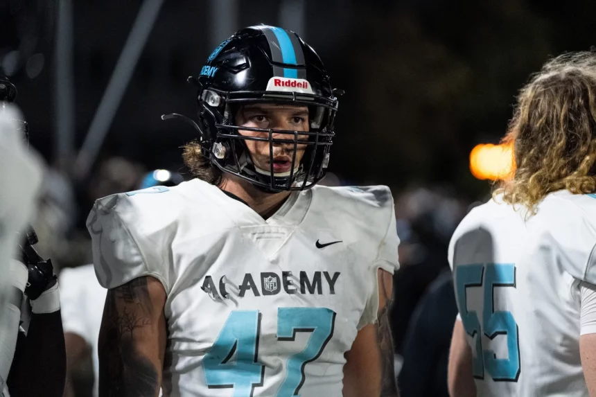 Auf dem Spielfeld steht Kevin Bentin mit schwarzem Helm und weißem Trikot mit der Nummer 47 und „ACADEMY“ auf der Vorderseite. Unter dem Helm sind lange Haare des Spielers zu sehen. Ein anderer Spieler, teilweise zu sehen, trägt ein Trikot mit der Nummer 55. Der Hintergrund ist schwach beleuchtet. Diese Beschreibung wurde mit der FootballR KI automatisch generiert.