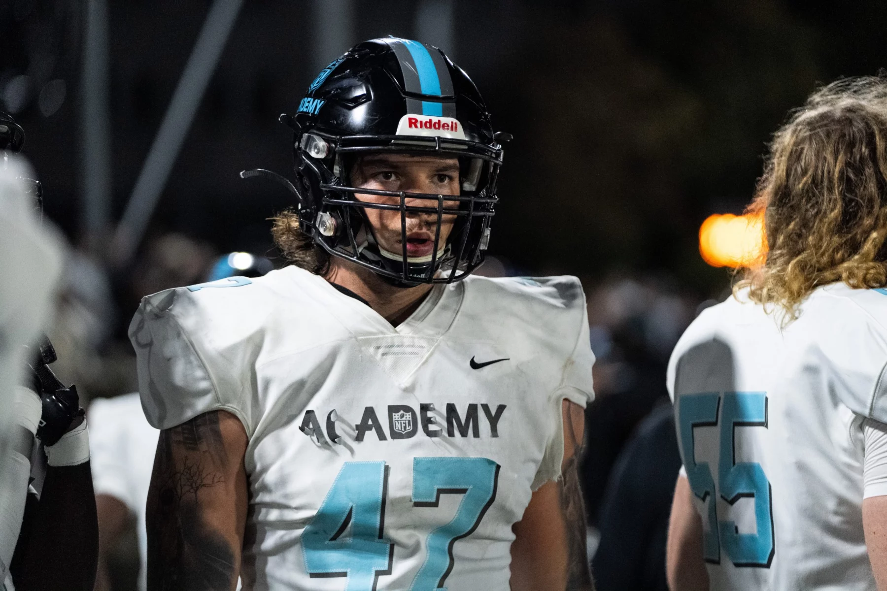 Auf dem Spielfeld steht Kevin Bentin mit schwarzem Helm und weißem Trikot mit der Nummer 47 und „ACADEMY“ auf der Vorderseite. Unter dem Helm sind lange Haare des Spielers zu sehen. Ein anderer Spieler, teilweise zu sehen, trägt ein Trikot mit der Nummer 55. Der Hintergrund ist schwach beleuchtet. Diese Beschreibung wurde mit der FootballR KI automatisch generiert.