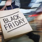 Black Friday Game: Eine Person hält eine große Einkaufstüte mit der Aufschrift „BLACK FRIDAY“ in fetten Buchstaben in der Hand und schlängelt sich durch das geschäftige Einkaufszentrum, ganz so, als würden Fans das spannende NFL-Spiel Raiders vs. Chiefs am Black Friday erwarten. Die verschwommenen Lichter und Displays fangen die lebhafte Popularität dieses Einkaufsrauschs ein. Diese Beschreibung wurde mit der FootballR KI automatisch generiert.
