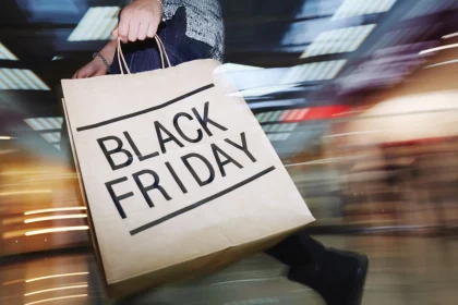 Black Friday Game: Eine Person hält eine große Einkaufstüte mit der Aufschrift „BLACK FRIDAY“ in fetten Buchstaben in der Hand und schlängelt sich durch das geschäftige Einkaufszentrum, ganz so, als würden Fans das spannende NFL-Spiel Raiders vs. Chiefs am Black Friday erwarten. Die verschwommenen Lichter und Displays fangen die lebhafte Popularität dieses Einkaufsrauschs ein. Diese Beschreibung wurde mit der FootballR KI automatisch generiert.