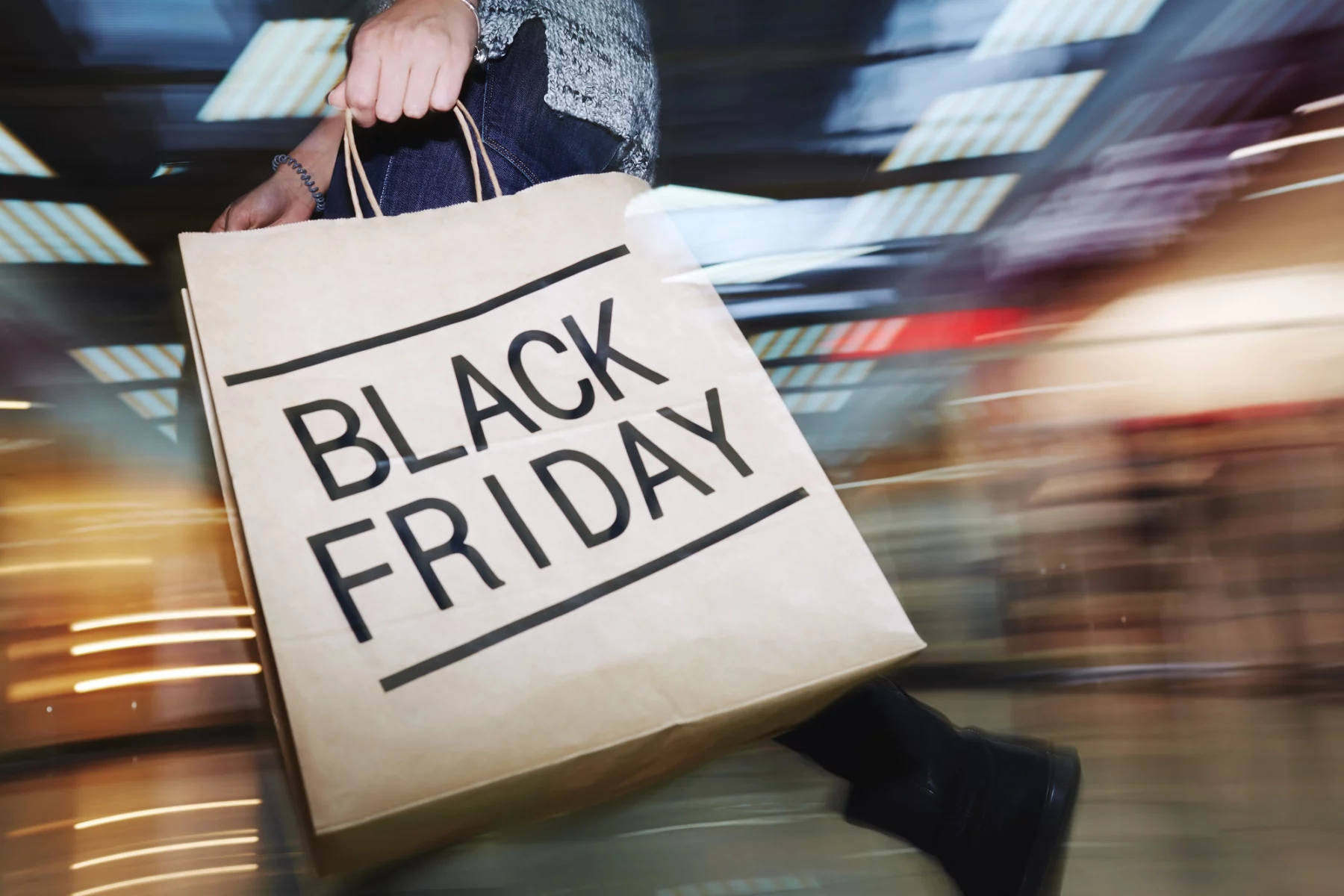 Black Friday Game: Eine Person hält eine große Einkaufstüte mit der Aufschrift „BLACK FRIDAY“ in fetten Buchstaben in der Hand und schlängelt sich durch das geschäftige Einkaufszentrum, ganz so, als würden Fans das spannende NFL-Spiel Raiders vs. Chiefs am Black Friday erwarten. Die verschwommenen Lichter und Displays fangen die lebhafte Popularität dieses Einkaufsrauschs ein. Diese Beschreibung wurde mit der FootballR KI automatisch generiert.