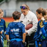 Ein Trainer mit einem Football gibt einer Gruppe von Kindern Anweisungen, die blaue Sporttrikots mit „NFL Flag“-Logos tragen, die mit der NFL Deutschland in Verbindung stehen. Die Kinder hören während einer Trainingseinheit auf einem Rasenplatz aufmerksam zu. Im Hintergrund ist eine Laufbahn zu sehen. Diese Beschreibung wurde mit der FootballR KI automatisch generiert.