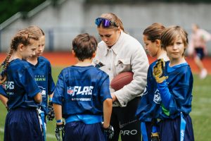 Ein Trainer mit einem Football gibt einer Gruppe von Kindern Anweisungen, die blaue Sporttrikots mit „NFL Flag“-Logos tragen, die mit der NFL Deutschland in Verbindung stehen. Die Kinder hören während einer Trainingseinheit auf einem Rasenplatz aufmerksam zu. Im Hintergrund ist eine Laufbahn zu sehen. Diese Beschreibung wurde mit der FootballR KI automatisch generiert.