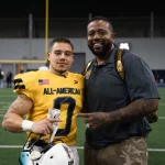Justus Seelig in einem gelben „All-American“-Trikot, das an den All-American Bowl der US Army erinnert, steht mit einem Helm in der Hand auf einem Feld. Neben ihm legt Ein Mann in Freizeitkleidung einen Arm um seine Schulter, während sie für ein Foto posieren, im Hintergrund sind Torpfosten zu sehen. Diese Beschreibung wurde mit der FootballR KI automatisch generiert.