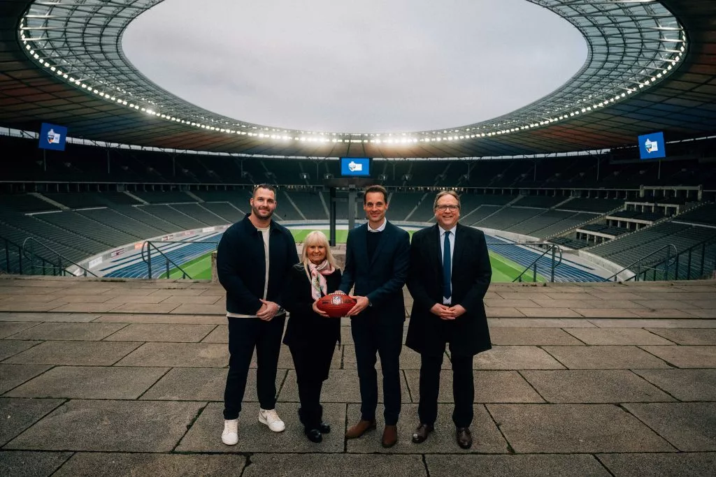 NFL Spielplan 2025 - NFL Berlin 2025: Vier Personen stehen in einem großen, leeren Stadion. Die zweite Person von rechts hält einen American Football. Die Gruppe trägt formelle Kleidung und das Stadion wird von Deckenlampen beleuchtet. Im Hintergrund sind das Spielfeld und die Sitzbereiche unter einem bewölkten Himmel zu sehen. Diese Beschreibung wurde mit der FootballR KI automatisch generiert.