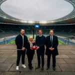 NFL Berlin 2025: Vier Personen stehen in einem großen, leeren Stadion. Die zweite Person von rechts hält einen American Football. Die Gruppe trägt formelle Kleidung und das Stadion wird von Deckenlampen beleuchtet. Im Hintergrund sind das Spielfeld und die Sitzbereiche unter einem bewölkten Himmel zu sehen. Diese Beschreibung wurde mit der FootballR KI automatisch generiert.