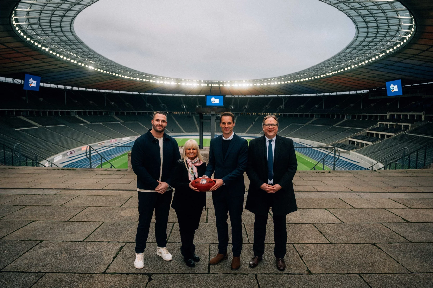 NFL Berlin 2025: Vier Personen stehen in einem großen, leeren Stadion. Die zweite Person von rechts hält einen American Football. Die Gruppe trägt formelle Kleidung und das Stadion wird von Deckenlampen beleuchtet. Im Hintergrund sind das Spielfeld und die Sitzbereiche unter einem bewölkten Himmel zu sehen. Diese Beschreibung wurde mit der FootballR KI automatisch generiert.
