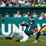 RTL NFL: Ein Footballspieler im blaugrünen Trikot (Nummer 1) gleitet gekonnt über das Feld und hält dabei den Ball. Ein anderer Spieler im weiß-schwarzen Trikot (Nummer 51) weicht einem Verteidiger der RTL NFL im schwarz-goldenen Trikot (Nummer 34) aus und stürmt auf ihn zu. Die Zuschauer auf den Tribünen dahinter beobachten ihn aufmerksam. Diese Beschreibung wurde mit der FootballR KI automatisch generiert.