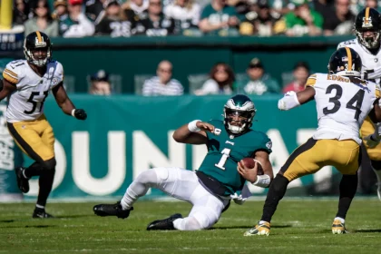 RTL NFL: Ein Footballspieler im blaugrünen Trikot (Nummer 1) gleitet gekonnt über das Feld und hält dabei den Ball. Ein anderer Spieler im weiß-schwarzen Trikot (Nummer 51) weicht einem Verteidiger der RTL NFL im schwarz-goldenen Trikot (Nummer 34) aus und stürmt auf ihn zu. Die Zuschauer auf den Tribünen dahinter beobachten ihn aufmerksam. Diese Beschreibung wurde mit der FootballR KI automatisch generiert.