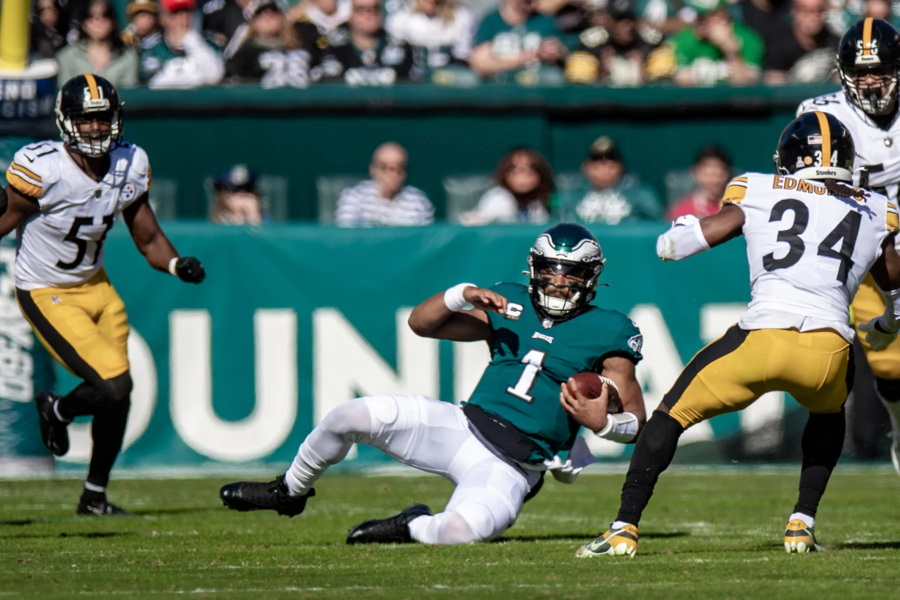 RTL NFL: Ein Footballspieler im blaugrünen Trikot (Nummer 1) gleitet gekonnt über das Feld und hält dabei den Ball. Ein anderer Spieler im weiß-schwarzen Trikot (Nummer 51) weicht einem Verteidiger der RTL NFL im schwarz-goldenen Trikot (Nummer 34) aus und stürmt auf ihn zu. Die Zuschauer auf den Tribünen dahinter beobachten ihn aufmerksam. Diese Beschreibung wurde mit der FootballR KI automatisch generiert.